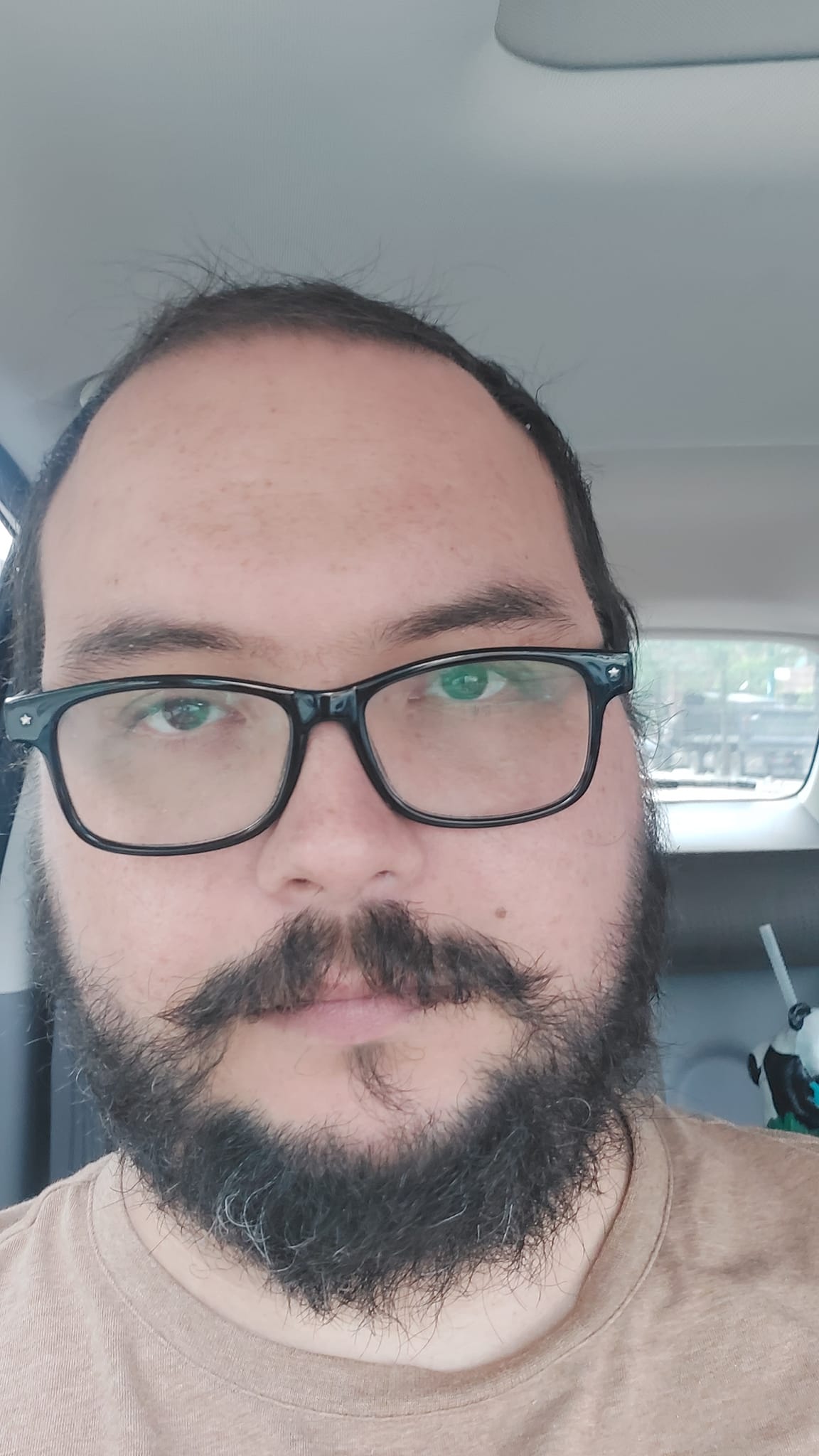 A man short hair and bushy facial hair stands sits in a car looking tired